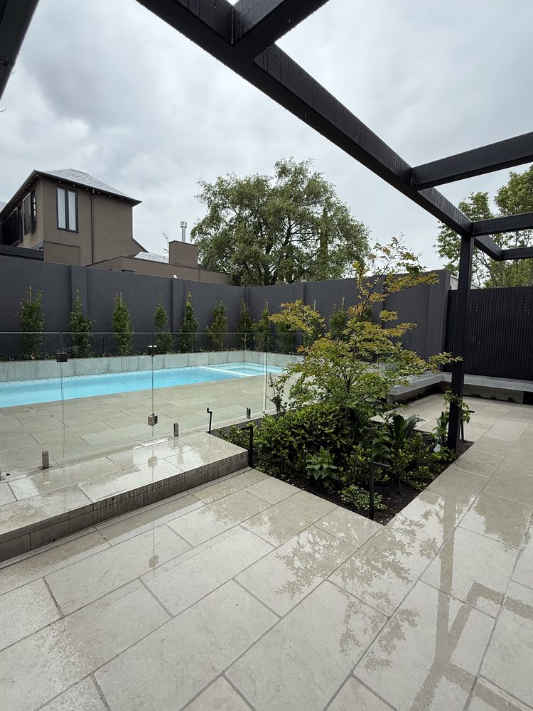 Pool with a frameless glass fence and limestone paving beside a planted garden bed