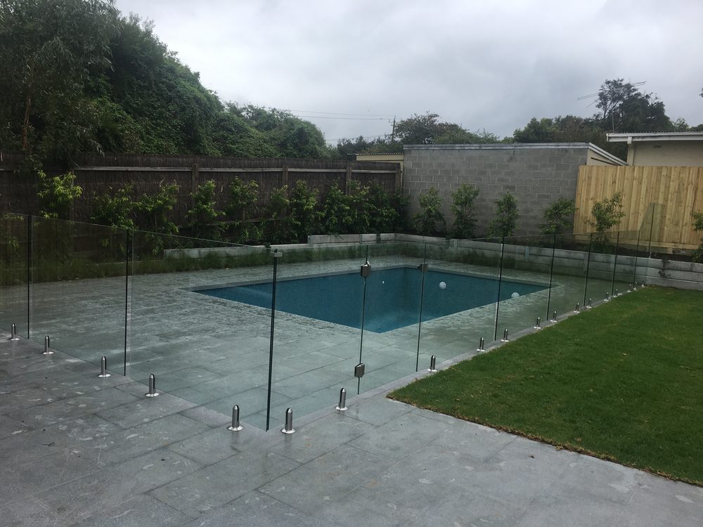 Frameless glass pool fence around a backyard pool with stone paving