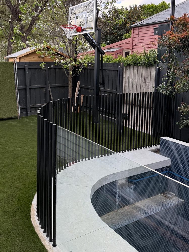 Curved black bar pool fence beside a tiled pool with a water blade and basketball hoop